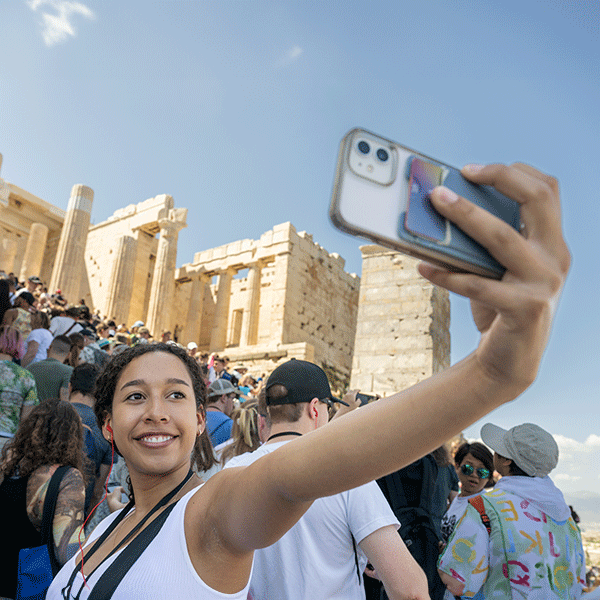 A smiling young woman of color wearing a white tank top takes a selfie with the Greek Parthenon in the background. The hand holding her phone is pictured in the foreground, and the Parthenon is visible at a high angle on a hill behind her amidst crowds of people.